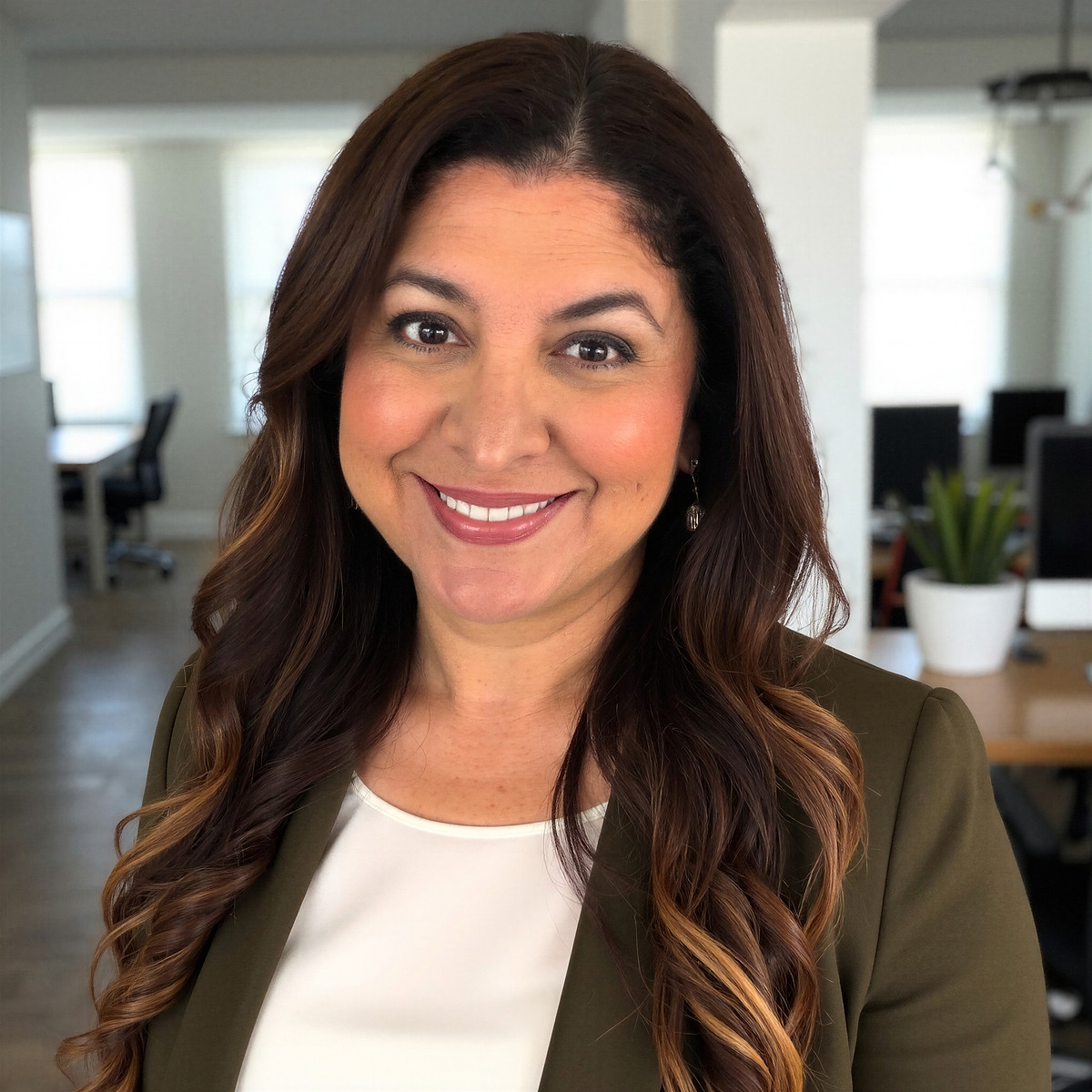Smiling woman with long brown hair and olive blazer in a bright open-office setting with desks and a plant in the background.